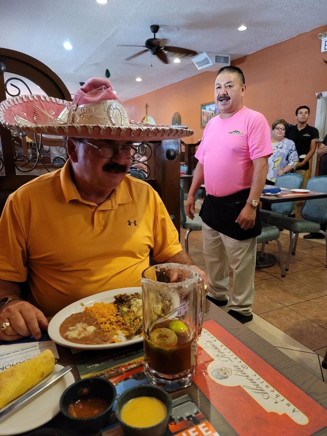 A guest wearing the restaurant's silver-trimmed sombrero at his table for his birthday, a server standing beside him, his plate and a tall drink in front of him