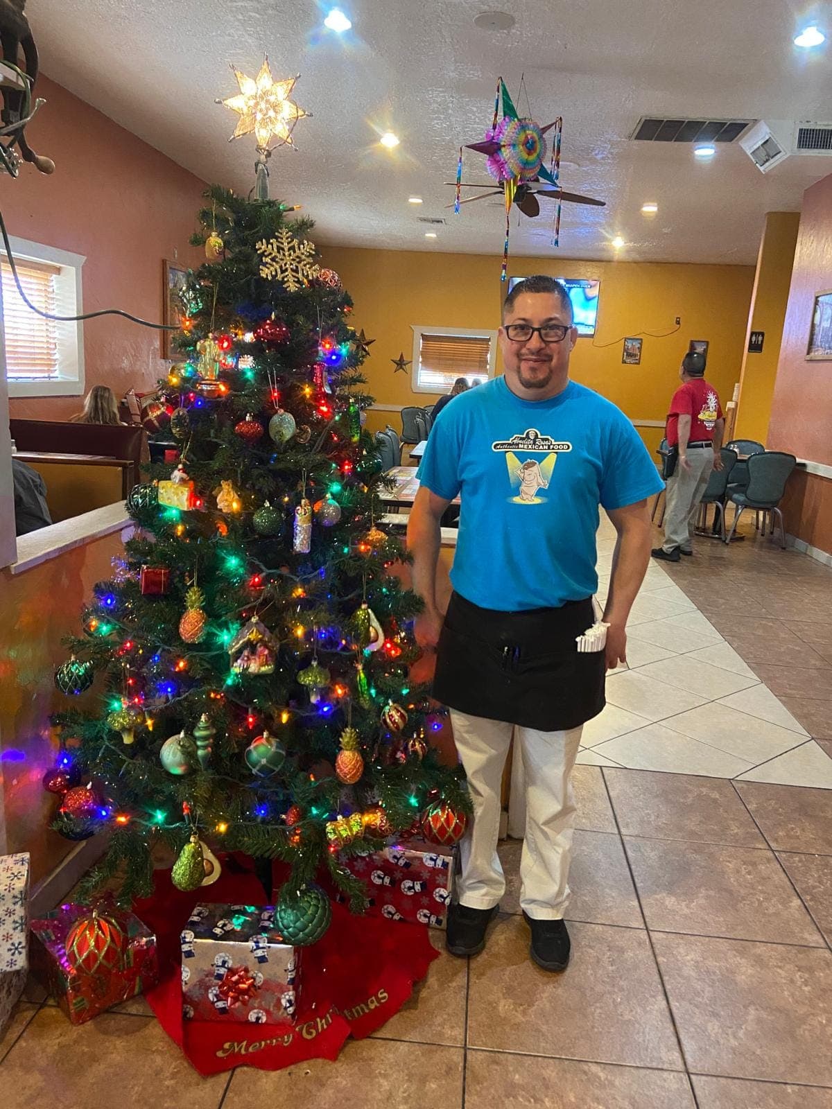 A server at Abuelita Rosa's standing beside the decorated Christmas tree in the dining room, smiling at the camera