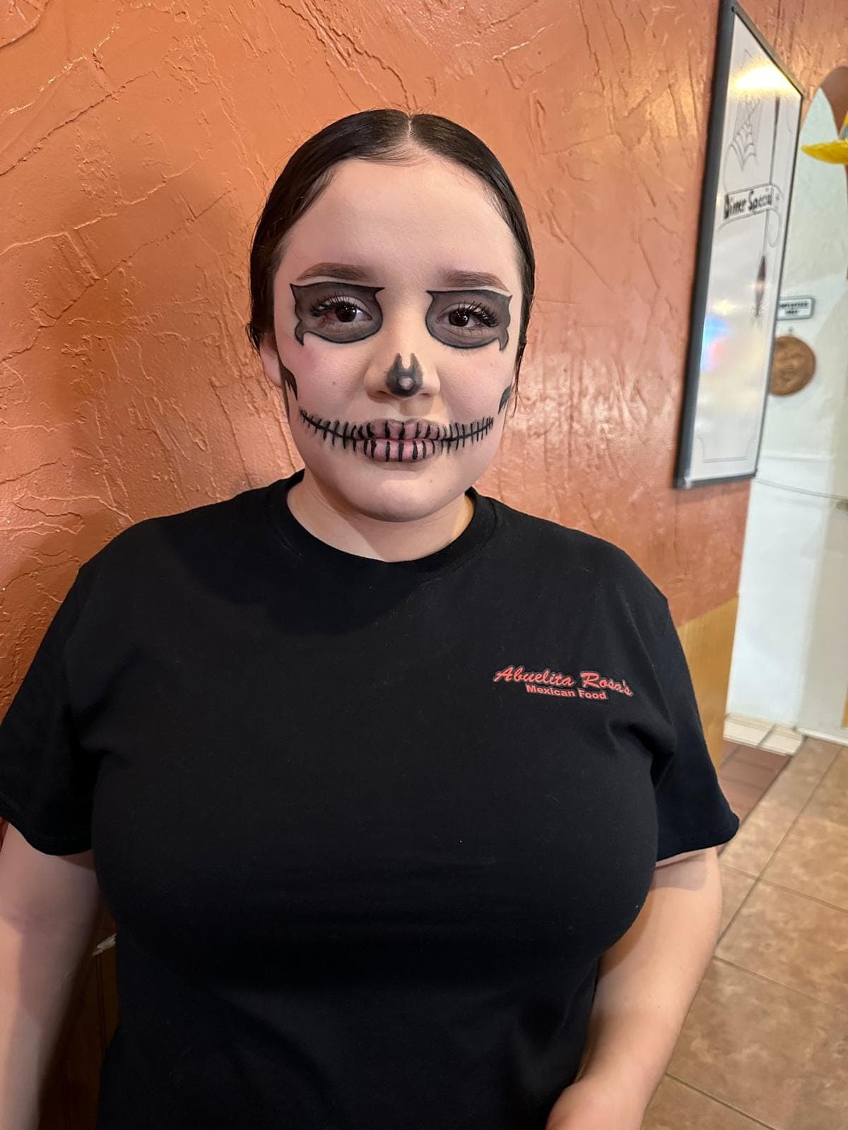 A server in an Abuelita Rosa's shirt with skull face paint for Halloween