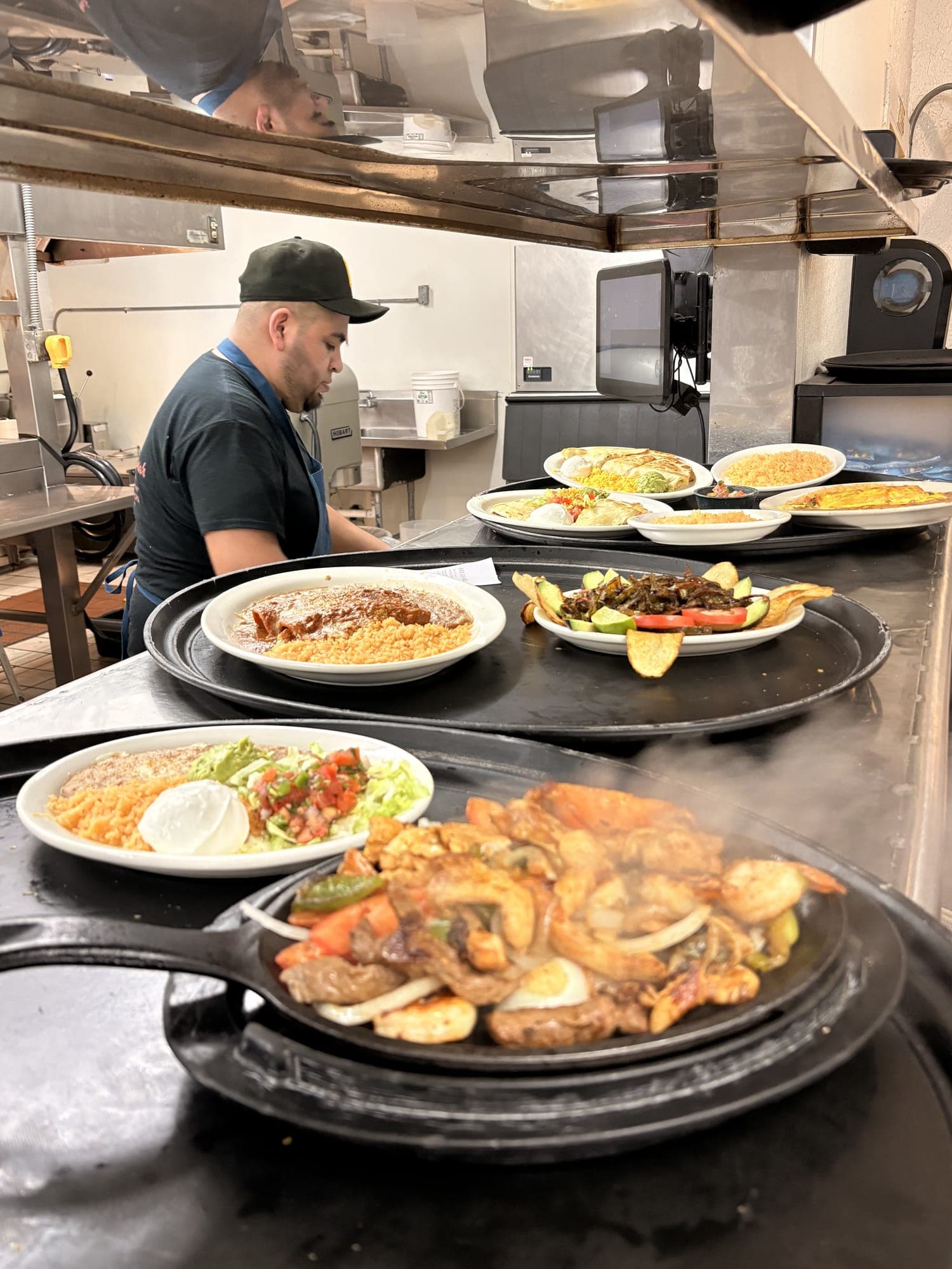 Sizzling fajitas on a cast iron skillet at the kitchen pass, with plates of enchiladas and rice waiting to go out